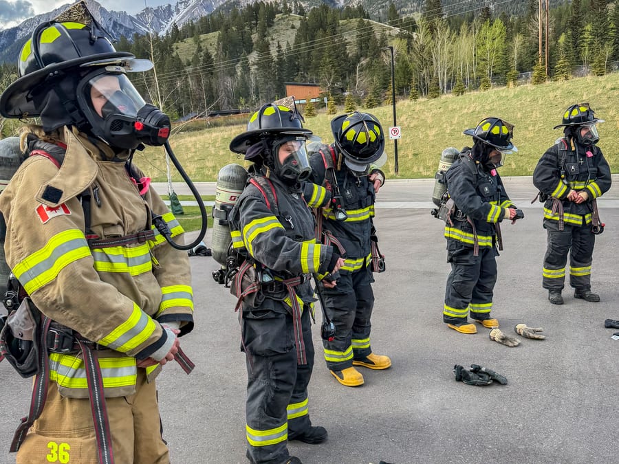 A line of firefighters in turnout gear and breathing apparatus waiting to begin a training evolution, with the Alberta Rockies behind them.