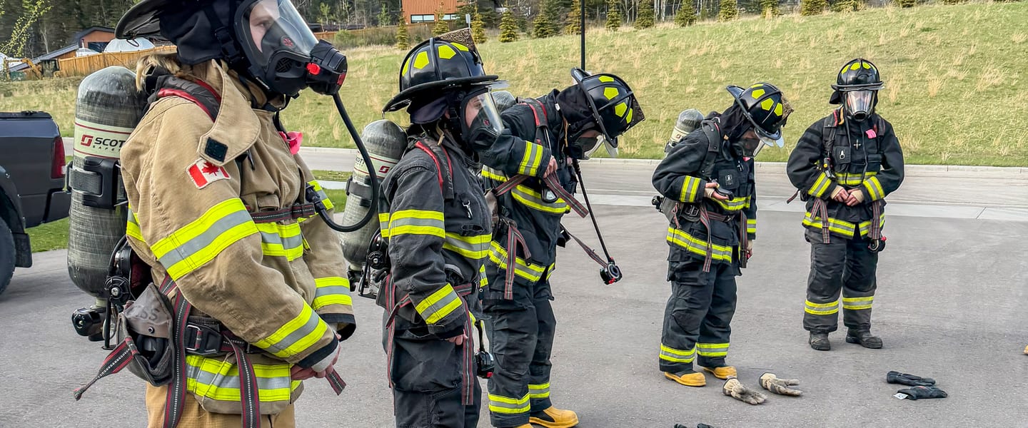 Firefighters in turnout gear and breathing apparatus on a training ground with the Rocky Mountains behind them.