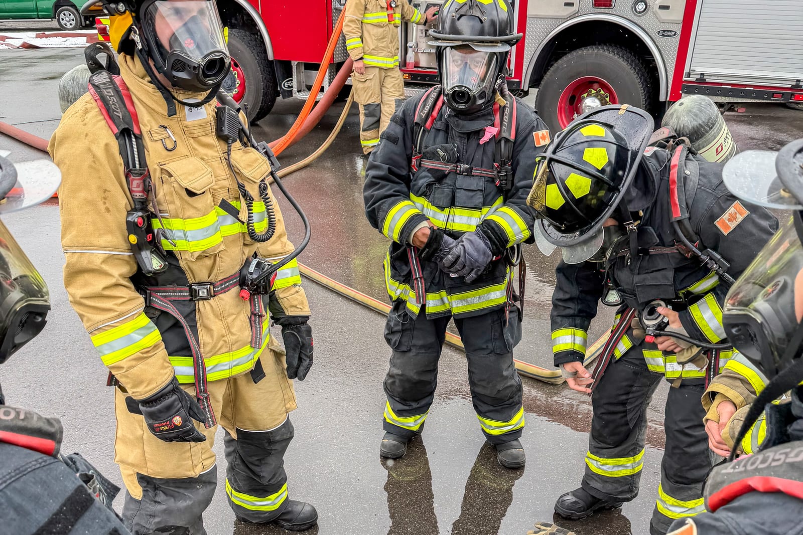 Firefighters in full turnout gear and breathing apparatus gathered beside a fire engine with hose lines deployed during a training exercise.
