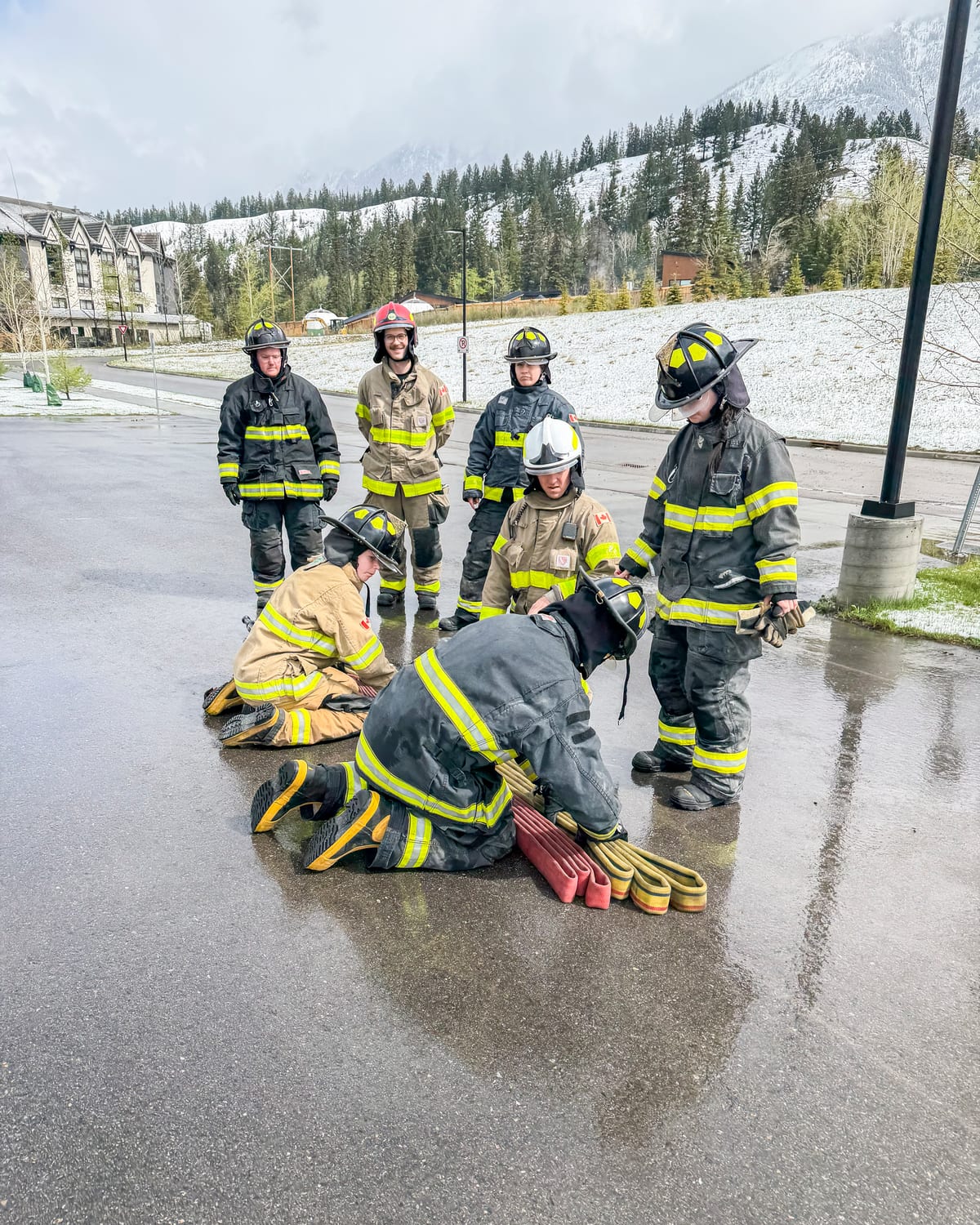 An instructor in a white helmet directing a hose evolution on a wet training ground while five firefighters observe.