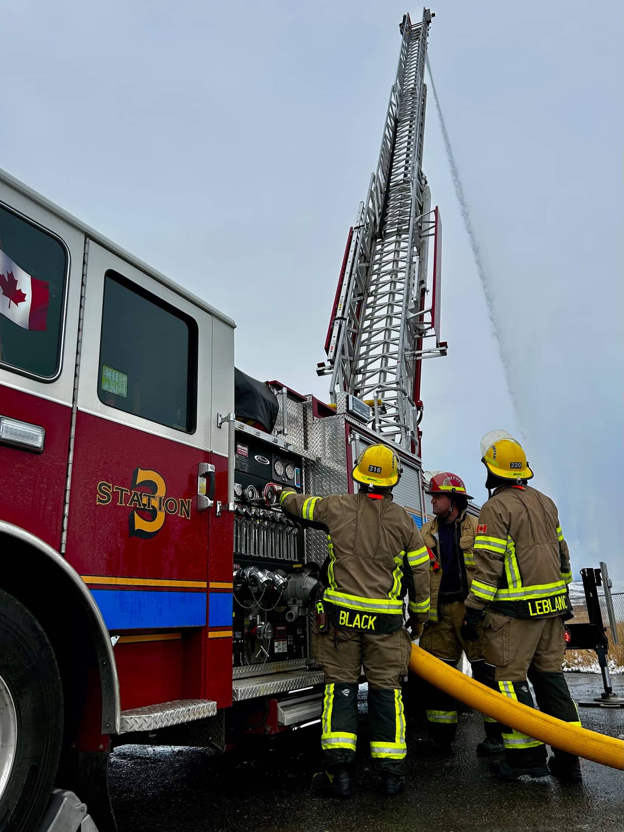 Three firefighters work the pump panel while a raised aerial ladder flows water overhead.