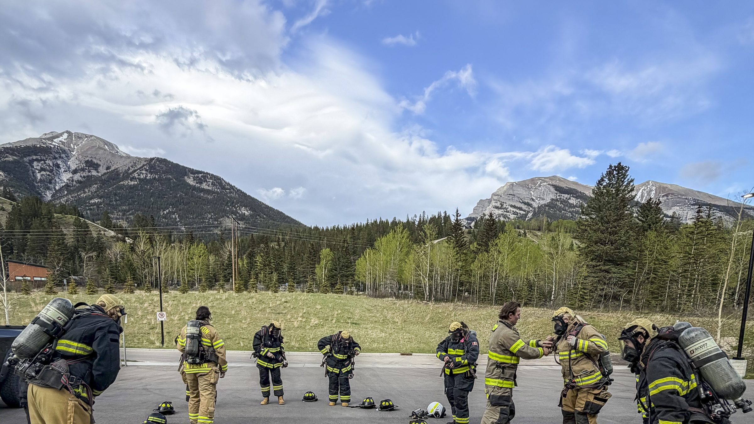 A crew in turnout gear donning SCBA on a paved training ground, with forested slopes and Rocky Mountain peaks behind them.