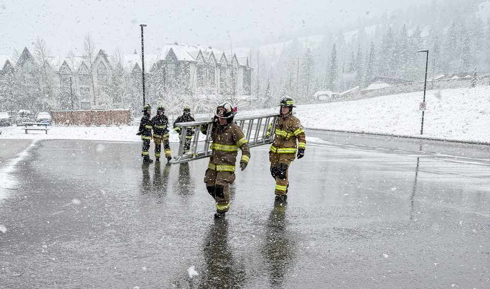 Two firefighters in turnout gear carrying a ladder across a wet, snow covered apron, with three more crew members behind them and a snow covered treeline beyond.