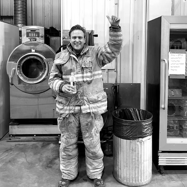 A firefighter in soiled turnout gear stands in a station equipment room, holding a drink and raising a thumb to the camera. Black and white photograph.