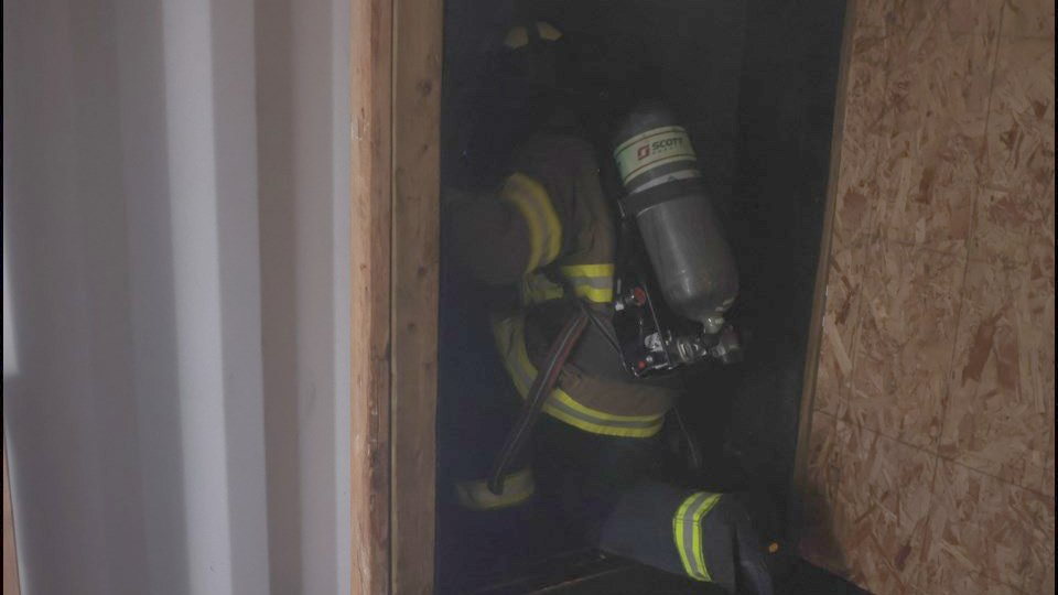 A firefighter on breathing apparatus moves through a narrow interior doorway in low light, cylinder and helmet outlined against the dark opening.