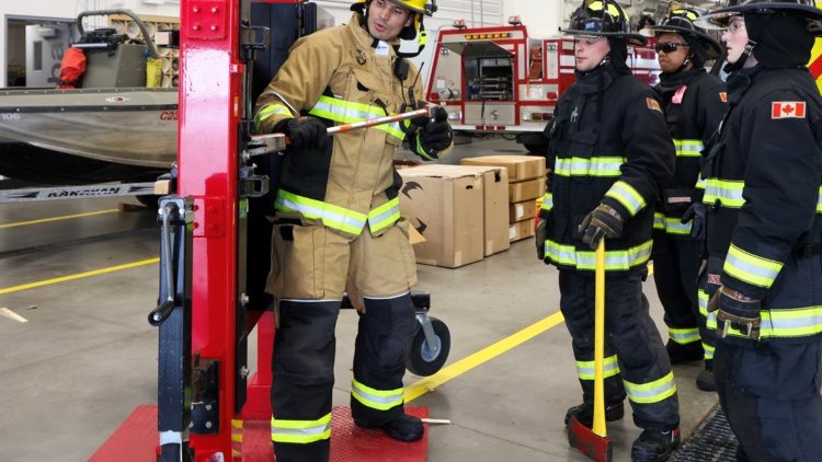 An instructor in turnout gear demonstrates with a hand tool at a red rescue strut inside an apparatus bay, five crew members in turnout gear standing and watching.