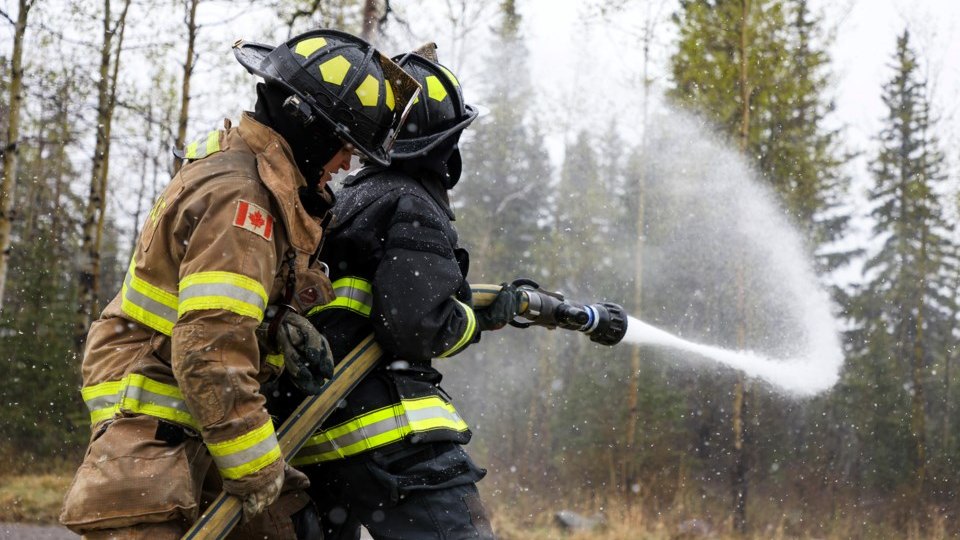 Two firefighters in turnout gear brace a charged hose line together, water streaming out in a wide arc, snow falling against evergreen trees behind them.