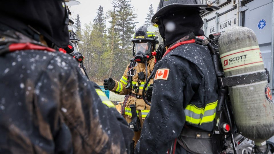 Two firefighters on self contained breathing apparatus stand in the foreground facing a third across a snow covered training ground, shipping containers behind them.