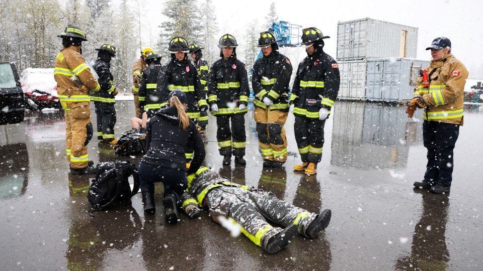 A line of firefighters in turnout gear watches while two crew members work over a patient lying on a snow covered lot, an instructor observing at the right.