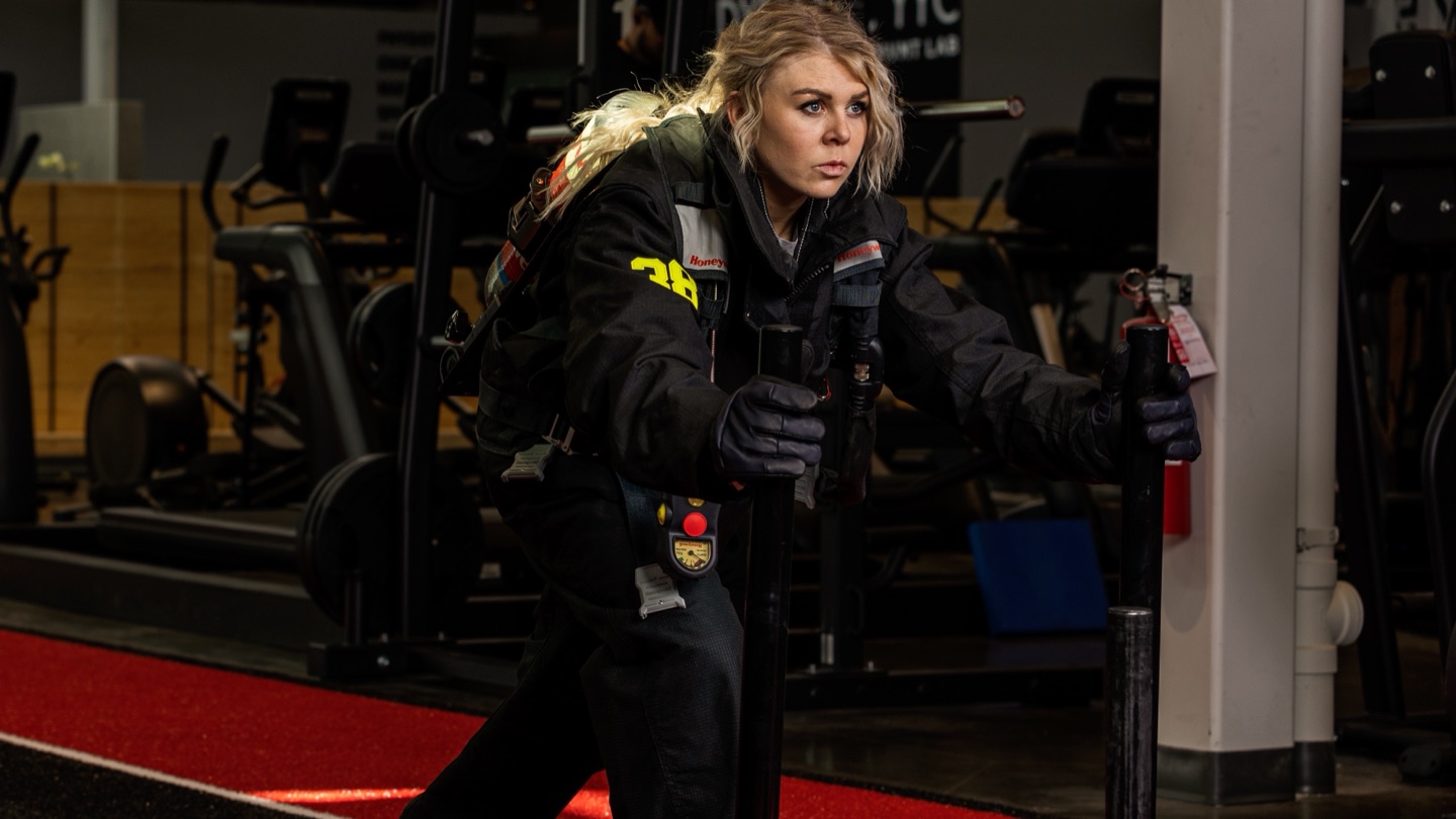 A firefighter in full turnout gear and breathing apparatus drives a weighted sled across a gym floor, leaning into the handles.