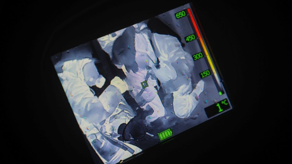 The screen of a thermal imaging camera showing several figures as white heat signatures against a dark background, with the temperature scale and battery indicator visible.